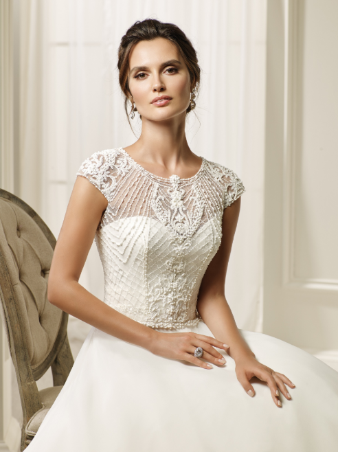 Close up of a brunette model sat on a plush grey chair in a beaded wedding dress with an embellished bodice, sweetheart illusion neckline and plain skirt.